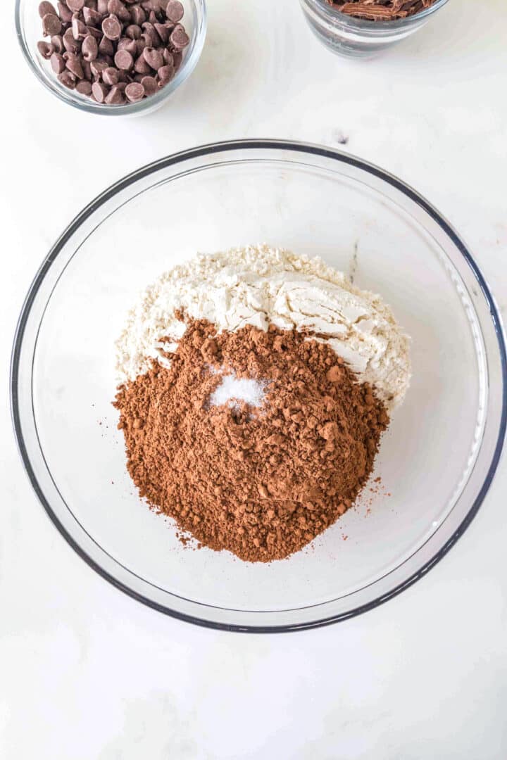 A glass bowl with flour, cocoa powder, and salt on a white surface, with small bowls of chocolate chips and chocolate shavings nearby.