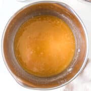 A metal mixing bowl filled with a light brown liquid mixture sits on a white surface, surrounded by bowls of chocolate shavings and cocoa powder.