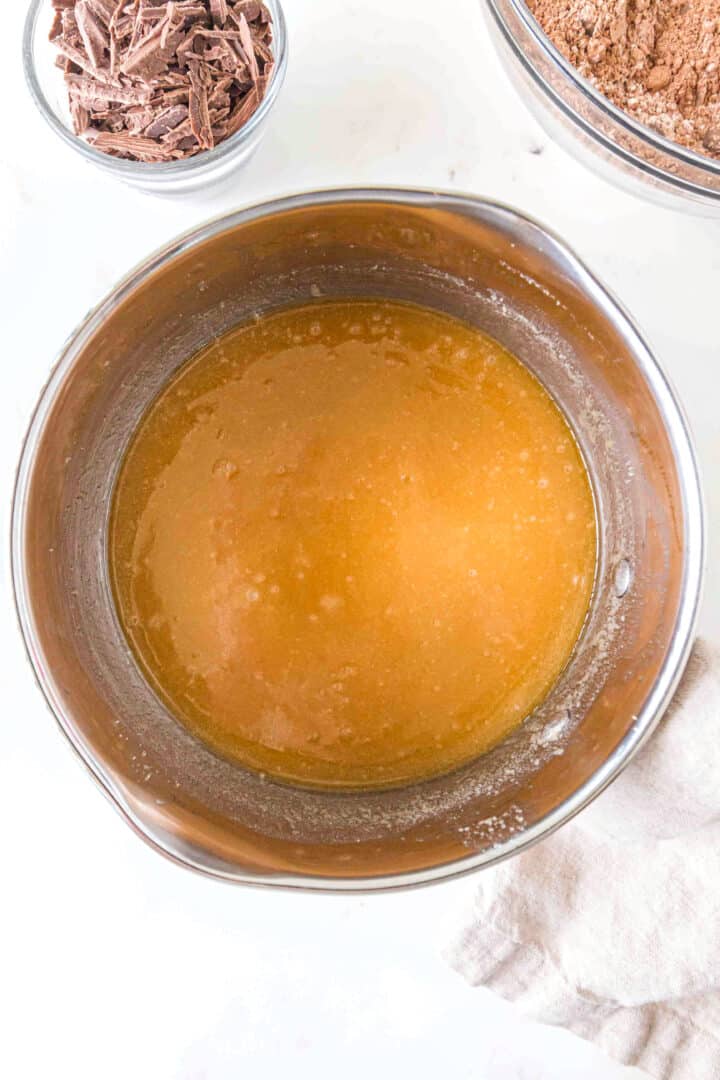 A metal mixing bowl filled with a light brown liquid mixture sits on a white surface, surrounded by bowls of chocolate shavings and cocoa powder.