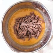 A metal mixing bowl containing a yellow batter with a pile of chopped chocolate pieces on top, ready to be mixed.