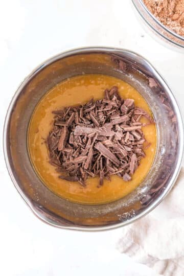 A metal mixing bowl containing a yellow batter with a pile of chopped chocolate pieces on top, ready to be mixed.