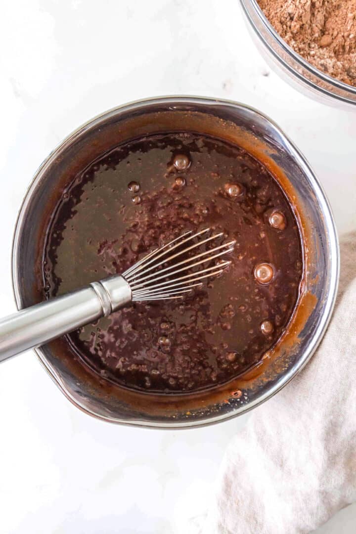 A metal whisk in a bowl of chocolate batter with visible bubbles, set on a light surface next to a beige cloth.