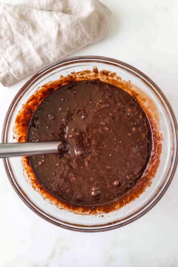 A glass bowl filled with chocolate cake batter being mixed with a metal whisk, placed on a white surface with a beige cloth nearby.