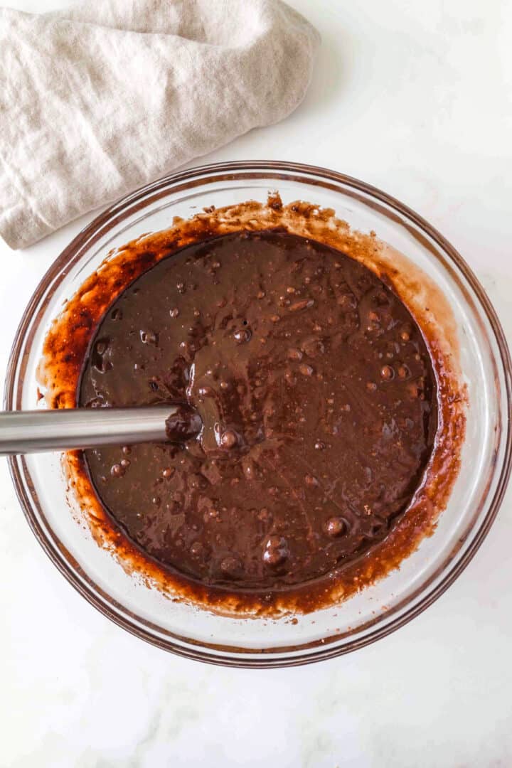 A glass bowl filled with chocolate cake batter being mixed with a metal whisk, placed on a white surface with a beige cloth nearby.