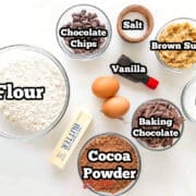 Top-down view of baking ingredients in bowls: flour, cocoa powder, sugar, brown sugar, chocolate chips, chocolate shavings, two eggs, butter, vanilla extract, and a small wooden bowl.