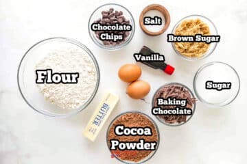 Top-down view of baking ingredients in bowls: flour, cocoa powder, sugar, brown sugar, chocolate chips, chocolate shavings, two eggs, butter, vanilla extract, and a small wooden bowl.