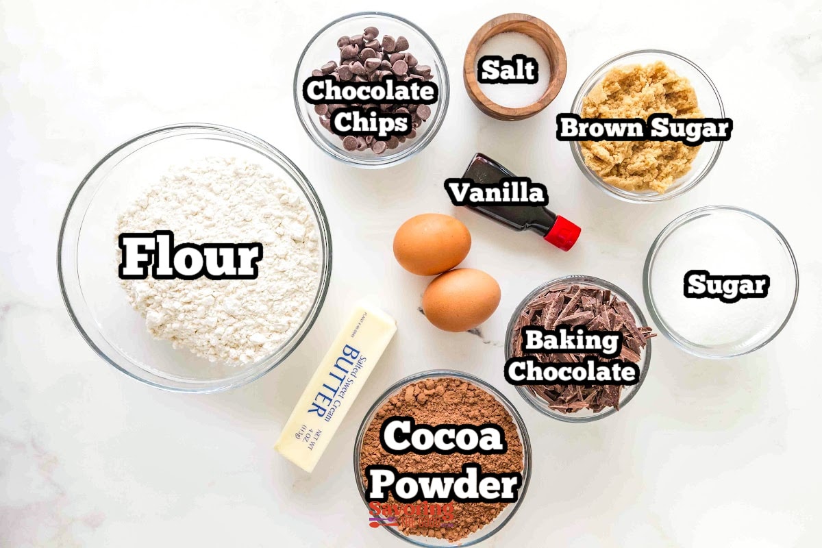 Top-down view of baking ingredients in bowls: flour, cocoa powder, sugar, brown sugar, chocolate chips, chocolate shavings, two eggs, butter, vanilla extract, and a small wooden bowl.