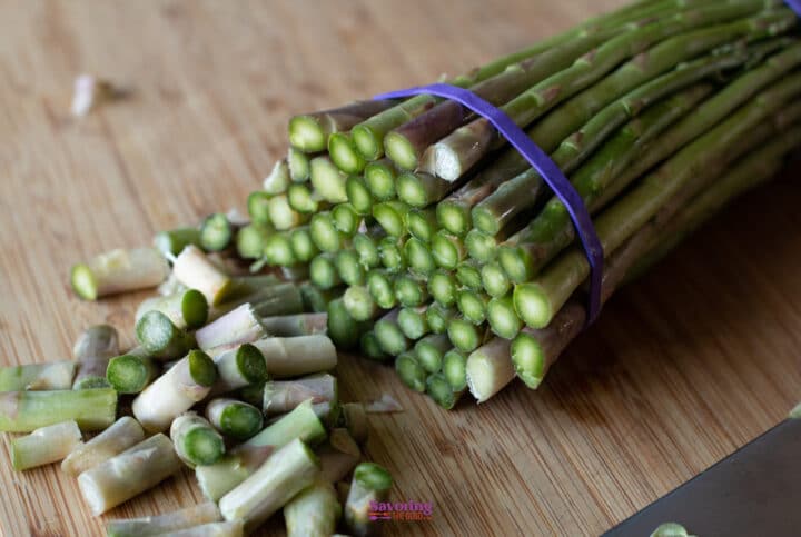 A bundle of fresh asparagus with trimmed ends sits on a wooden cutting board next to a pile of discarded stalk pieces.