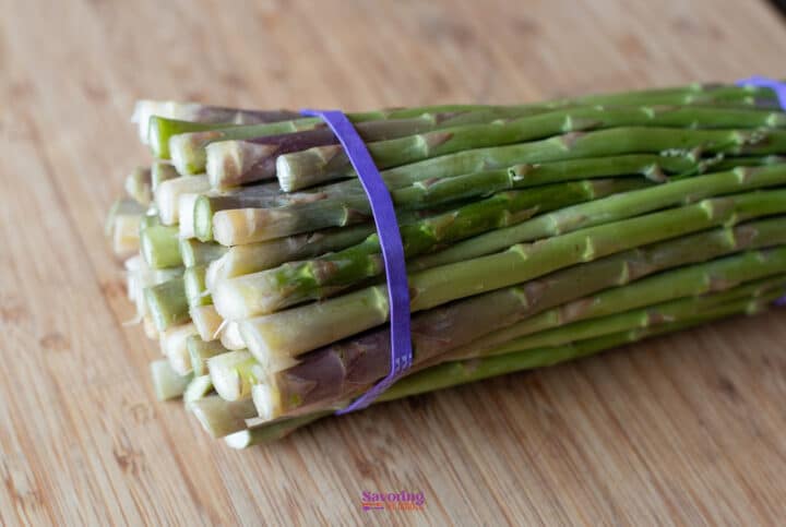 A bundle of fresh asparagus spears held together with a purple rubber band, placed on a wooden cutting board.