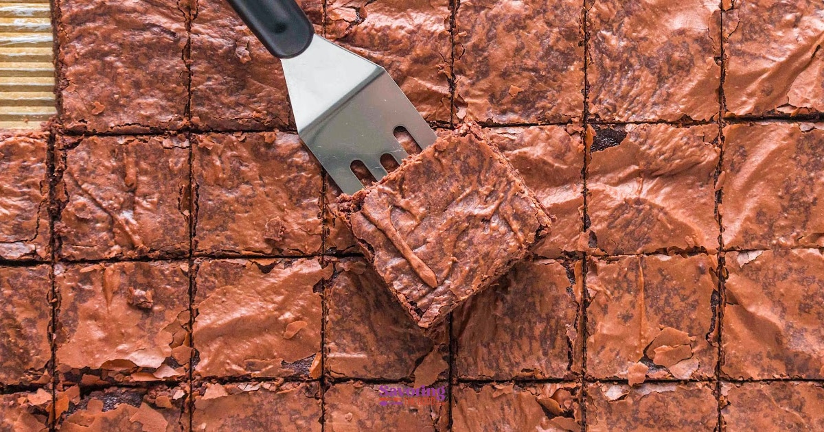 A spatula lifts a square brownie from a tray of sheet pan brownies cut into equal pieces.