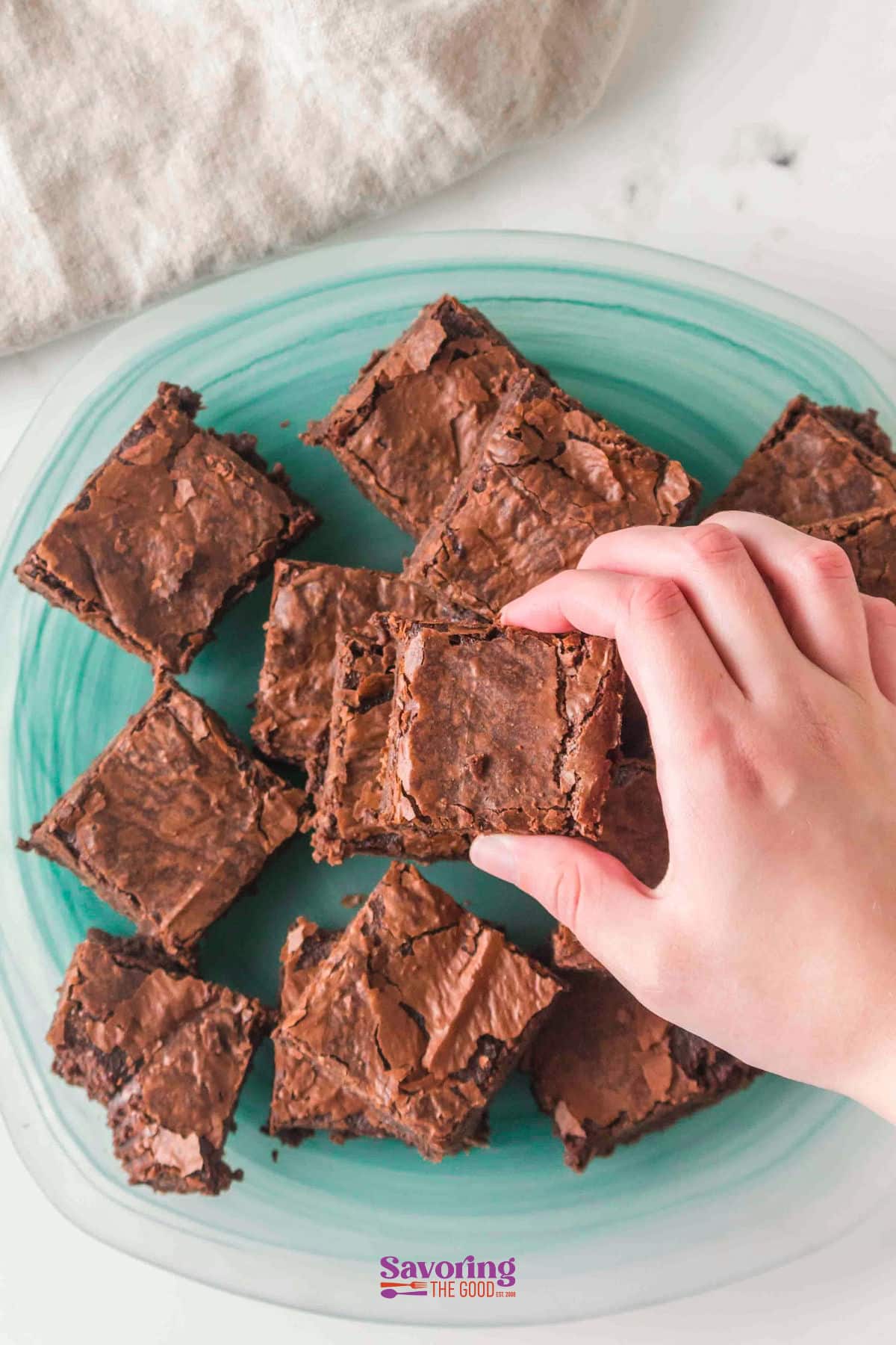 A hand reaches for a chocolate brownie from a plate with several sheet pan brownies arranged on a green oval dish.