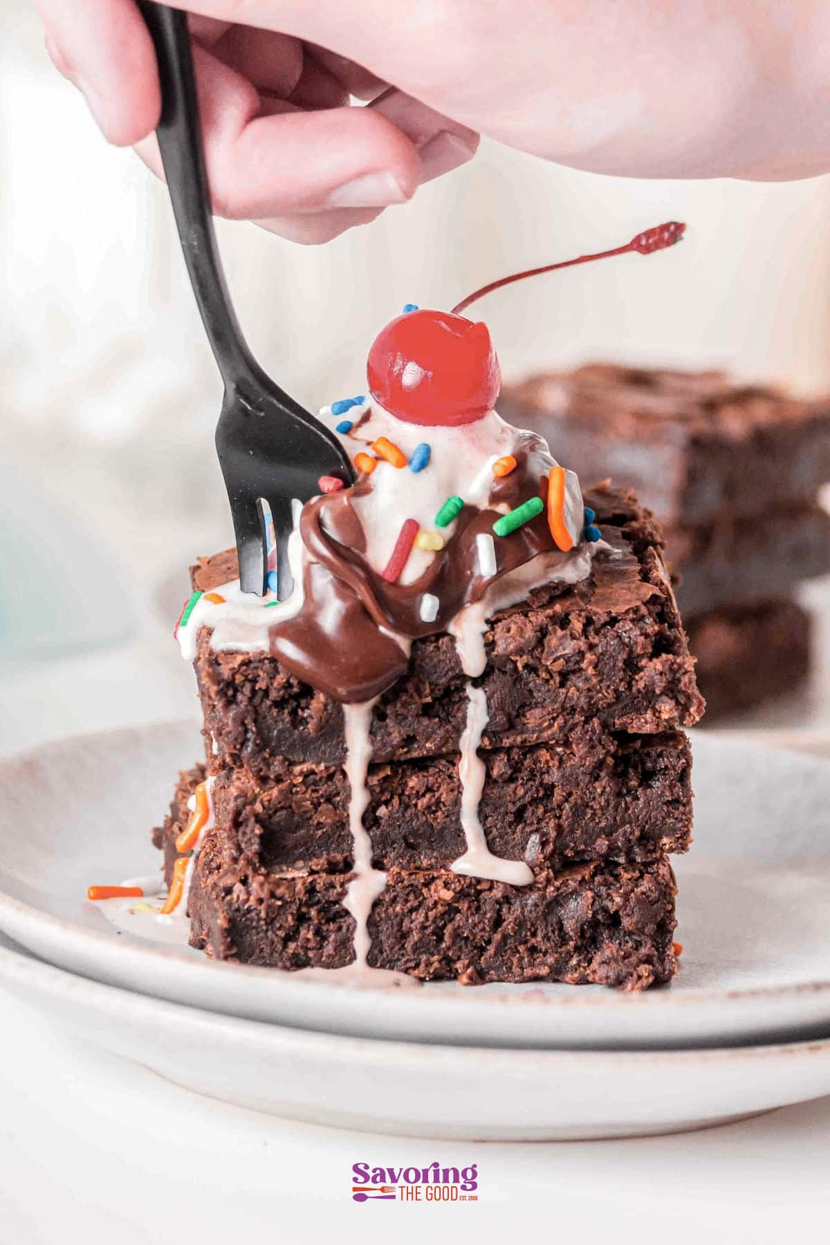 A hand using a fork to cut into a stack of sheet pan brownies topped with ice cream, chocolate sauce, sprinkles, and a cherry on a white plate.