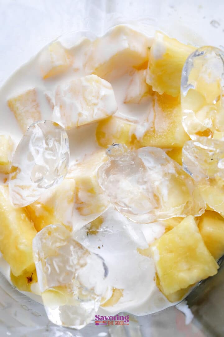 Chopped pineapple and ice cubes in a white liquid in a blender.
