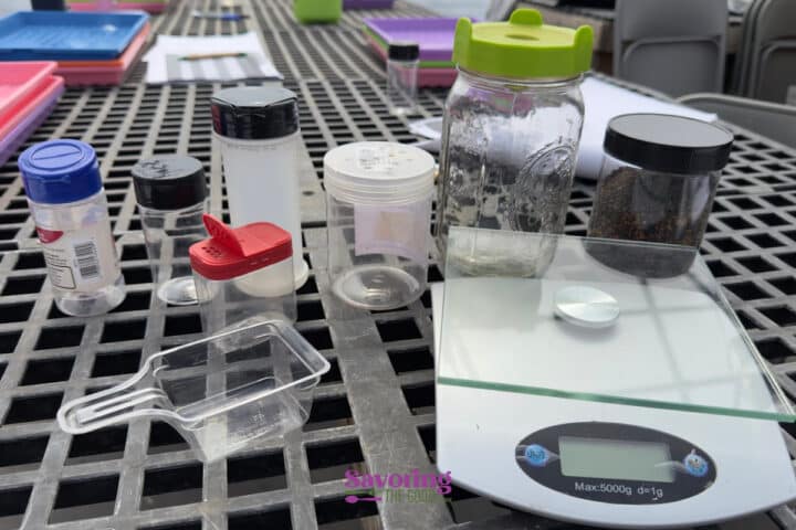 Assorted empty and partially filled plastic containers, a digital kitchen scale, and a clear scoop are arranged on a metal outdoor table.