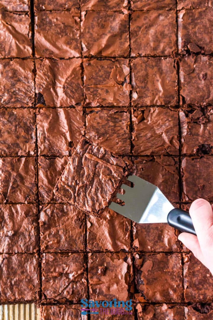 A hand holding a spatula lifts a square piece of chocolate brownie from a tray of evenly cut brownies.