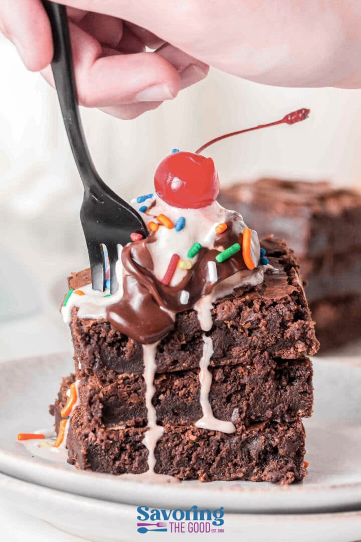 A hand uses a black fork to cut into a stack of brownies topped with ice cream, chocolate sauce, sprinkles, and a cherry.