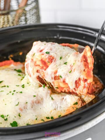 A serving of lazy lasagna is being lifted from a slow cooker with a metal spatula. The cheesy lasagna is topped with melted cheese and sprinkled parsley.