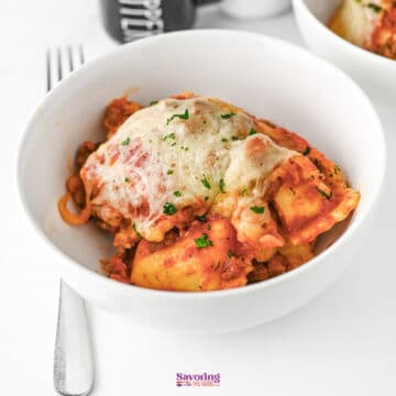 A white bowl filled with lazy lasagna, topped with melted cheese and herbs, sits on a white surface with a fork beside it.
