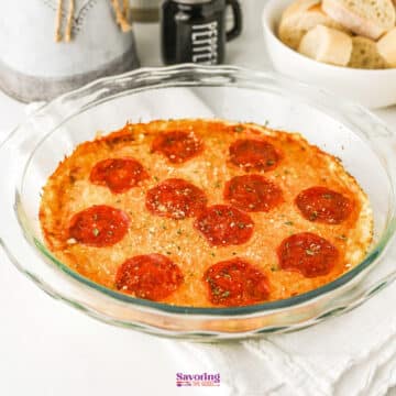 A glass baking dish filled with baked pizza dip featuring pepperoni and cheese, with a bowl of sliced bread in the background.