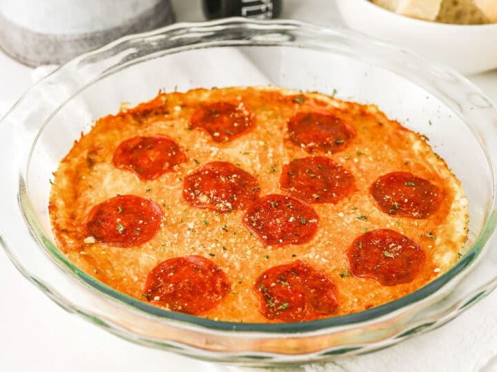 A glass baking dish filled with baked pizza dip featuring pepperoni and cheese, with a bowl of sliced bread in the background.