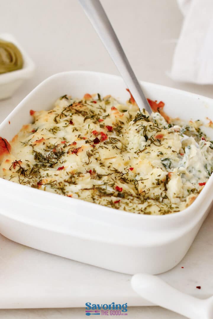 A white ceramic dish filled with baked, cheesy dip topped with herbs, with a spoon scooping some out.