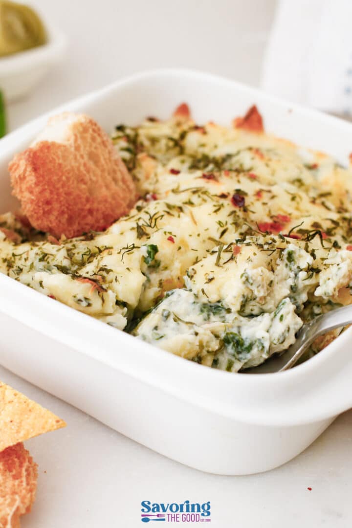 A white dish filled with creamy spinach artichoke dip, topped with herbs and served with a piece of bread and a metal spoon.