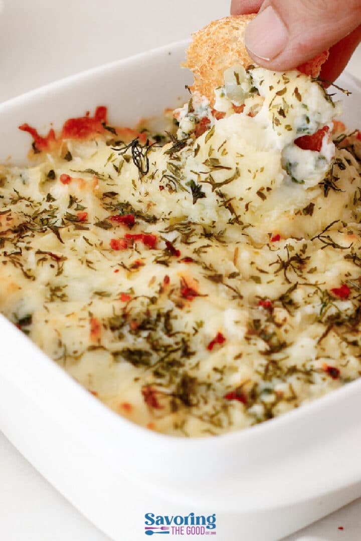 A hand dips bread into a baked, cheesy spinach and artichoke dip topped with herbs in a white square dish.