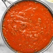 A skillet filled with red pasta sauce containing ground meat sits on a white cloth, with a small bowl of shredded cheese nearby.