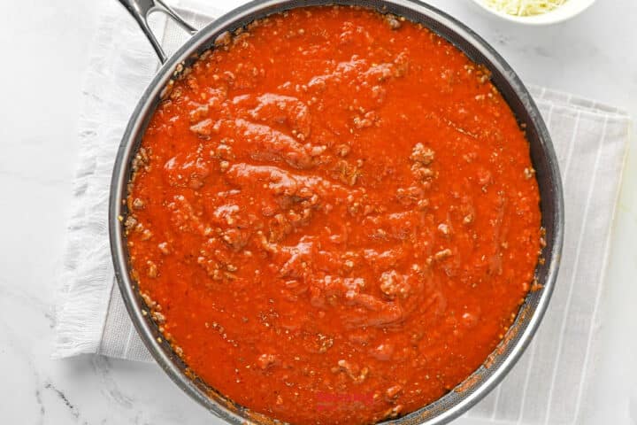 A skillet filled with red pasta sauce containing ground meat sits on a white cloth, with a small bowl of shredded cheese nearby.
