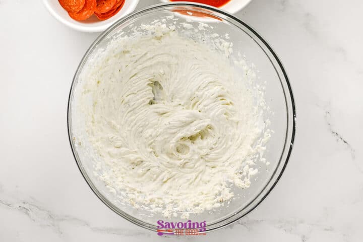 A glass bowl filled with whipped cream cheese mixture, perfect for pizza dip, sits on a marble surface, with bowls of pepperoni partially visible above.