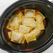 Uncooked ravioli placed in a black slow cooker over a layer of red pasta sauce, creating an easy lazy lasagna.
