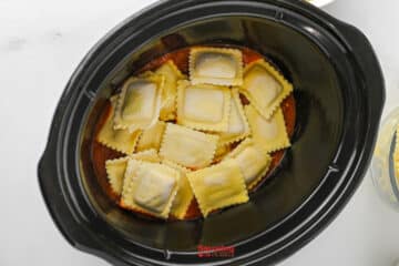 Uncooked ravioli placed in a black slow cooker over a layer of red pasta sauce, creating an easy lazy lasagna.