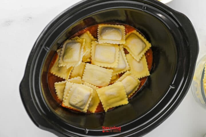 Uncooked ravioli placed in a black slow cooker over a layer of red pasta sauce, creating an easy lazy lasagna.