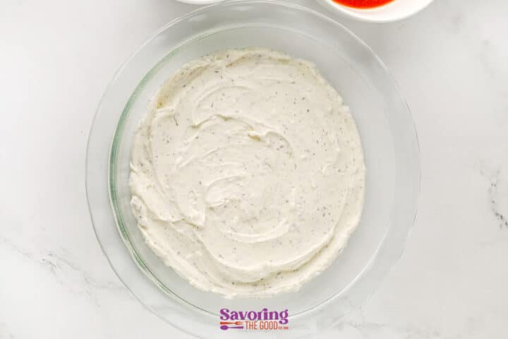 A glass pie dish filled with an even layer of creamy, white pizza dip, speckled with visible herbs mixed in.