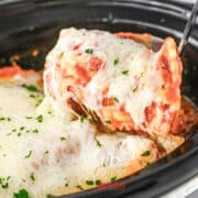 A serving of cheesy lasagna being lifted from a slow cooker, topped with melted cheese and garnished with parsley.