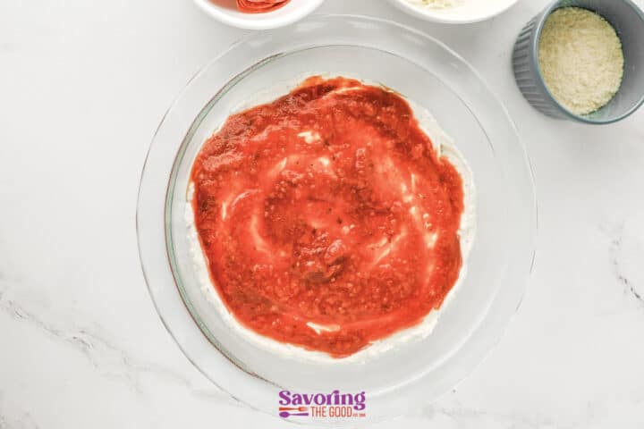 A glass pie dish with a layer of cream cheese topped with tomato sauce creates a delicious pizza dip; bowls of grated cheese are nearby on a marble surface.
