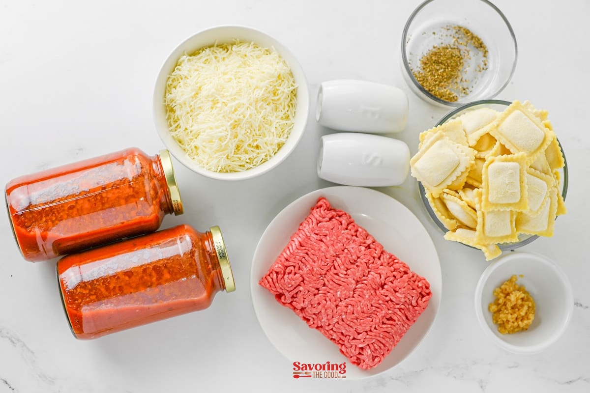 Top-down view of ingredients for a pasta dish, including ground beef, ravioli, shredded cheese, pasta sauce, minced garlic, seasoning, and salt and pepper shakers.
