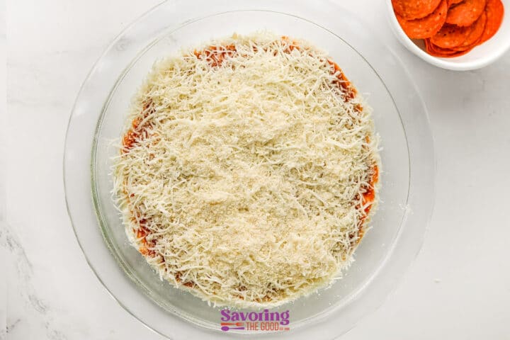 A glass pie dish filled with cheesy pizza dip sits next to a bowl of pepperoni slices on a white surface.