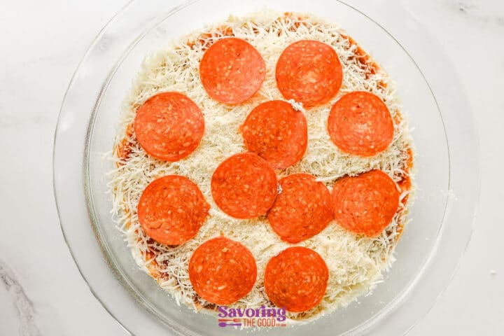 A glass dish filled with shredded cheese, pizza sauce, and a single layer of pepperoni slices arranged on top.