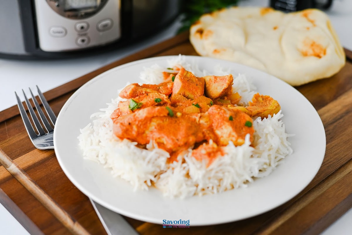 A plate of white rice topped with orange curry chicken, garnished with herbs, served with naan bread on a wooden tray beside a fork and knife. An Instant Pot in the background hints at slow cooker chicken tikka masala perfection.