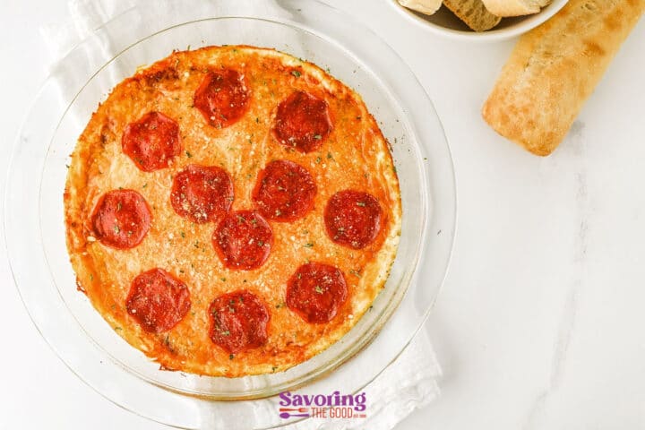 A round glass dish filled with baked dip topped with pepperoni slices, next to a piece of bread and a bowl of bread slices on a white surface.