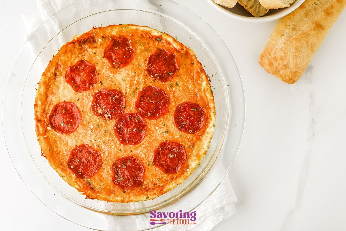 A round glass dish filled with baked dip topped with pepperoni slices, next to a piece of bread and a bowl of bread slices on a white surface.