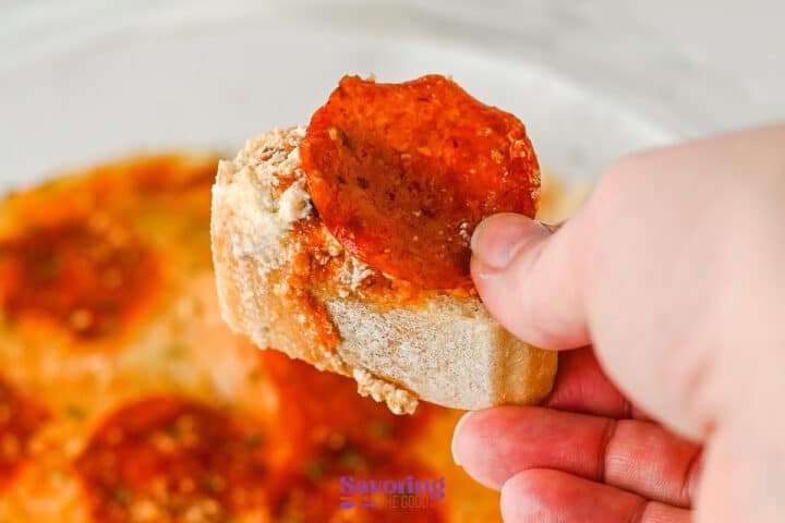 A hand holds a slice of bread topped with tomato sauce, grated cheese, and a pepperoni slice, resembling a pizza dip appetizer.