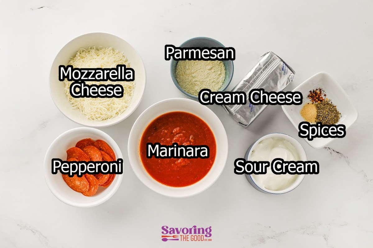Six bowls containing ingredients for a delicious pizza dip-shredded mozzarella cheese, pepperoni slices, marinara sauce, grated parmesan cheese, cream cheese, sour cream, and a small dish of spices-are arranged on a white surface.
