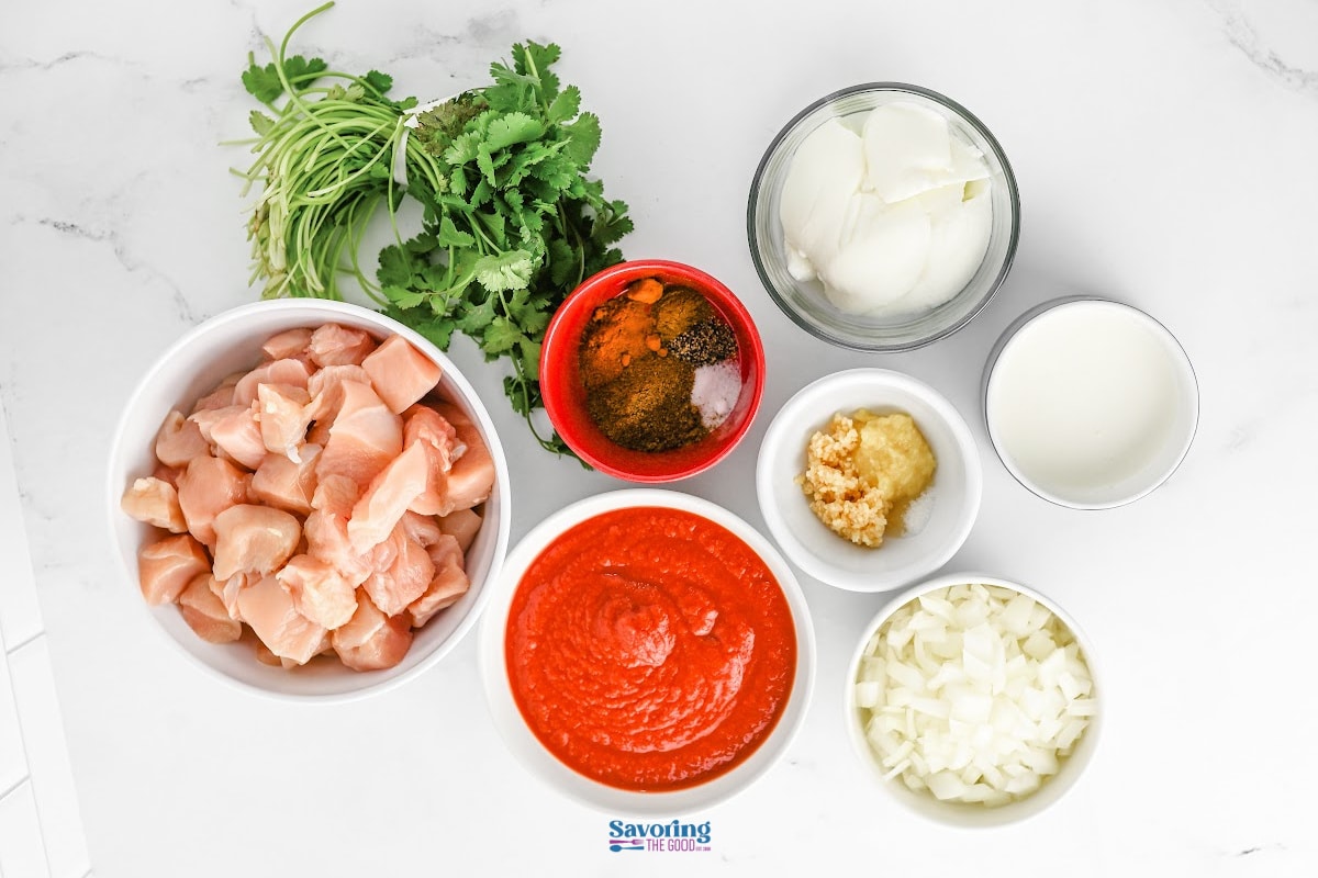 Overhead view of ingredients for slow cooker chicken tikka masala on a white surface: raw chicken pieces, fresh cilantro, spices, tomato sauce, chopped onions, minced garlic, sliced onions, and a bowl of cream.
