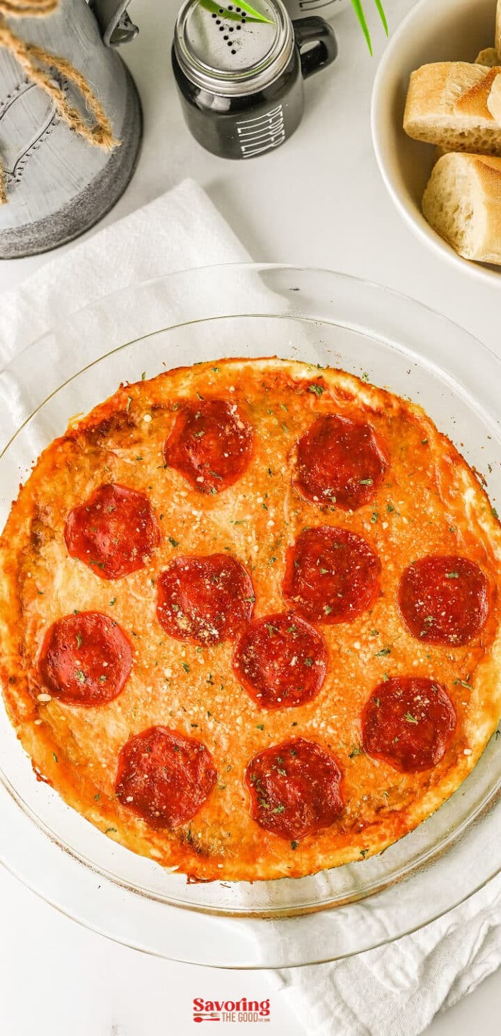 A glass pie dish filled with baked pepperoni pizza dip, topped with pepperoni slices and herbs, sits on a white cloth near a bowl of sliced bread.