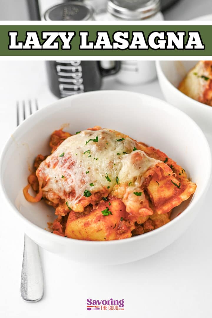 A bowl of lasagna topped with melted cheese, garnished with herbs, sits on a white surface with a fork and a pepper grinder in the background.