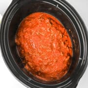 A slow cooker filled with red tomato sauce and ground meat, perfect for making lazy lasagna, sits on a white surface with a bowl of pasta nearby.