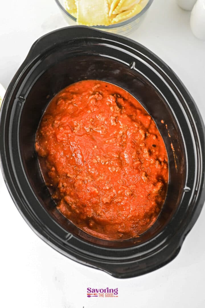 A slow cooker filled with red tomato sauce and ground meat, perfect for making lazy lasagna, sits on a white surface with a bowl of pasta nearby.