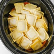 Uncooked ravioli arranged in a black slow cooker on top of a layer of tomato sauce, creating an easy lazy lasagna.